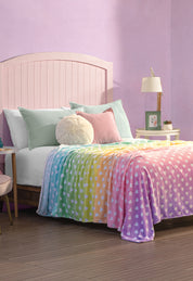 A cozy bedroom features a light pink headboard bed, pastel pillows, and the Cobertor Flannel Ligero Azúcar by Intima Hogar—a rainbow-colored blanket with white heart designs. Purple walls and nightstands with lamps, plants, and decor add extra charm.
