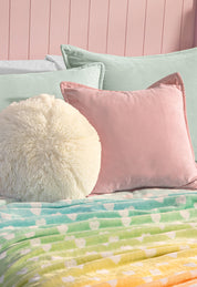 A cozy bed with pastel green and pink pillows, a round fluffy cream pillow, and the Intima Hogar Cobertor Flannel Ligero Azúcar—featuring a rainbow gradient and white heart design—set against a pink paneled wall.
