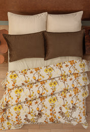 A neatly made bed with beige and brown pillows features the Intima Hogar Cobertor Flannel Extra Suave Colombia, displaying a yellow and brown floral design. The bed sits on a wooden floor with bedside tables on each side.