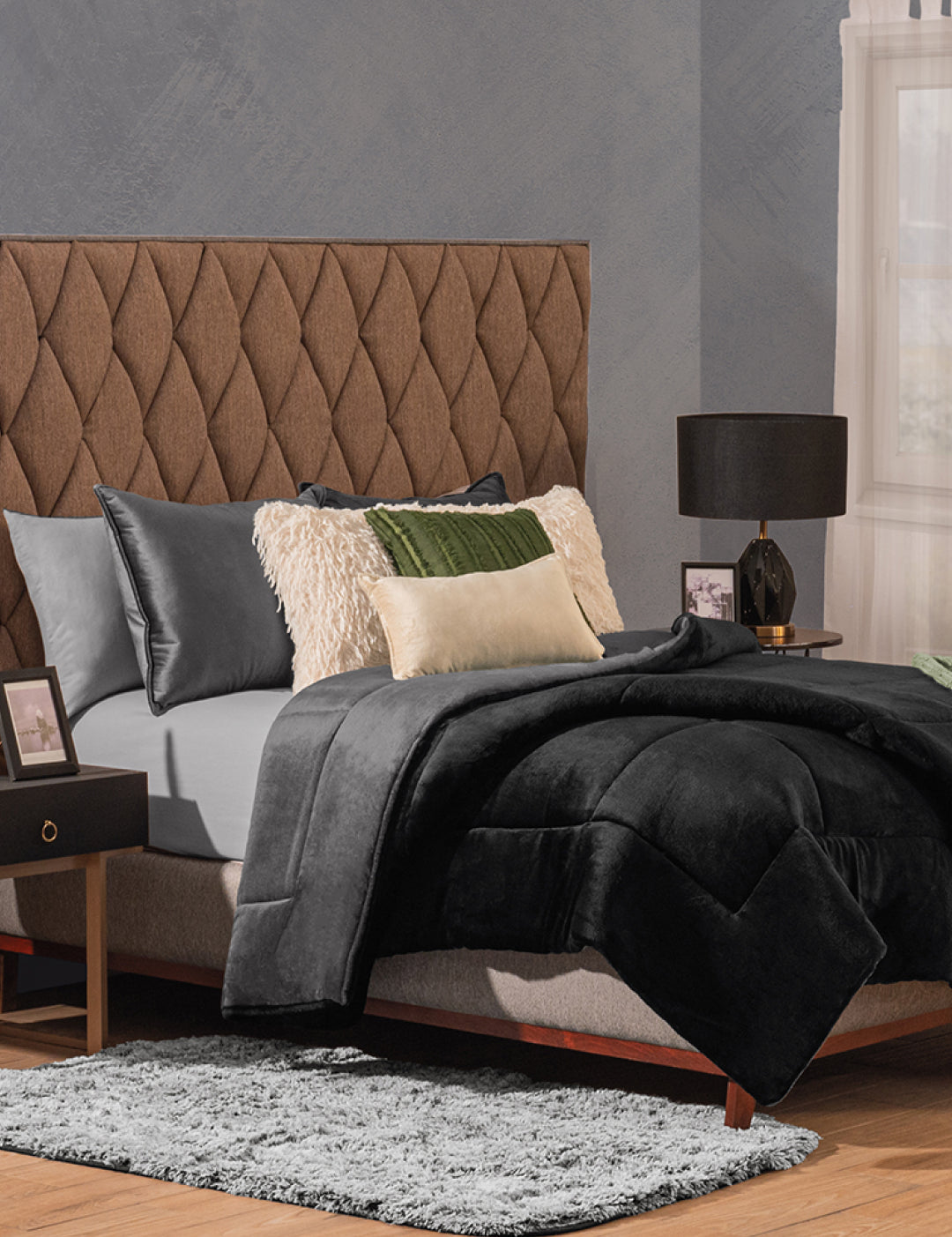 A modern bedroom with a brown tufted headboard, gray bedding, black and cream pillows, and the Cobertor Flannel Extra Suave Estambul by Intima Hogar for lasting warmth, plus a black bedside table with lamp and gray rug on wood floor near white-curtained window.