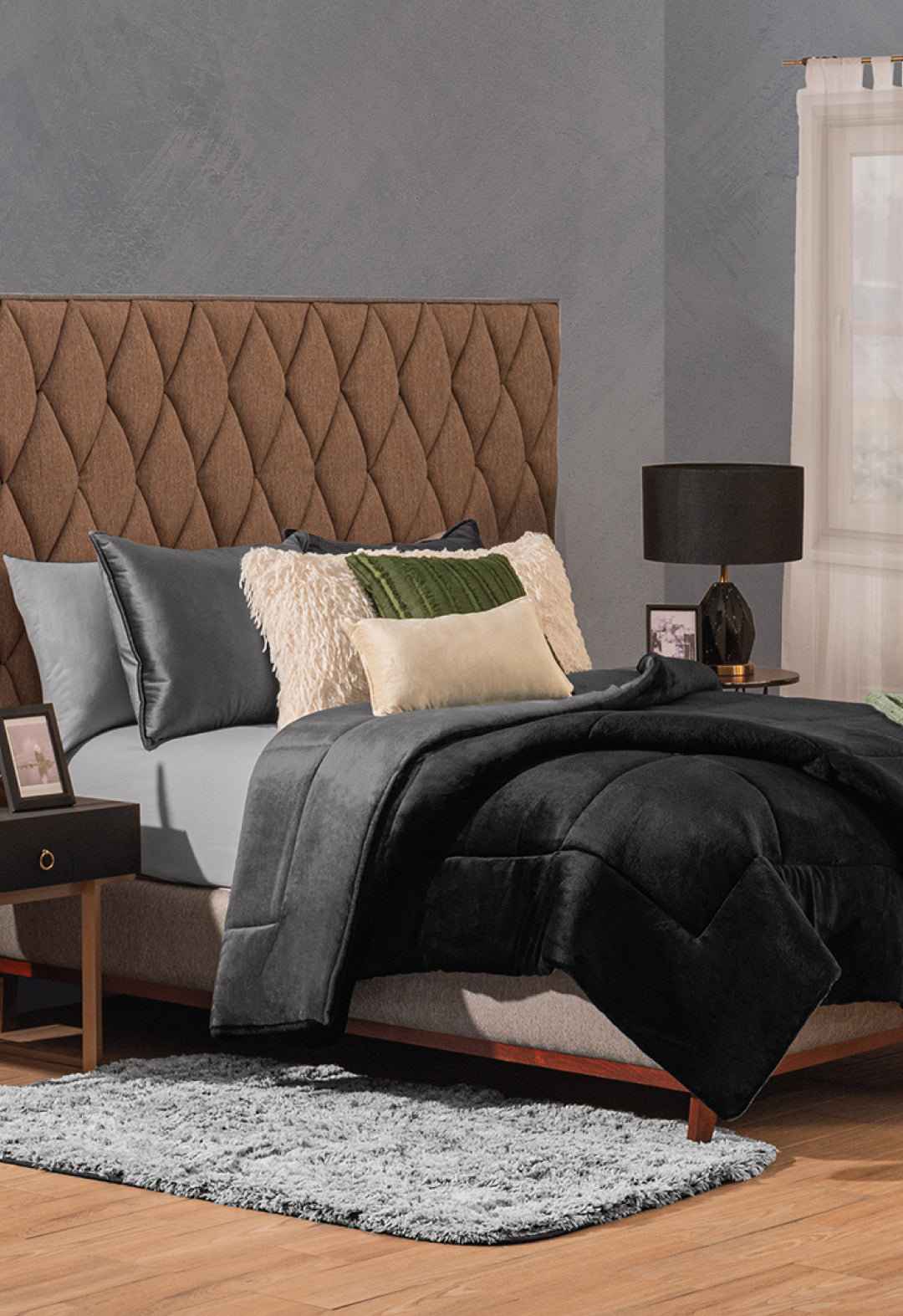 A modern bedroom with a brown tufted headboard, gray bedding, black and cream pillows, and the Cobertor Flannel Extra Suave Estambul by Intima Hogar for lasting warmth, plus a black bedside table with lamp and gray rug on wood floor near white-curtained window.