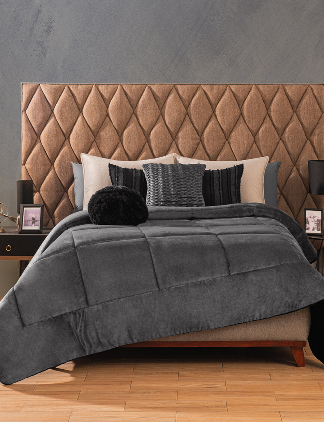 A neatly made bed features the Intima Hogar Cobertor Flannel Extra Suave Estambul in gray, paired with black and gray pillows and a tufted brown headboard. Two nightstands with lamps and framed photos sit against a textured gray wall.