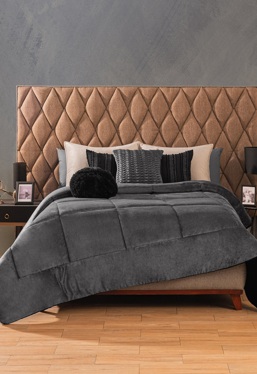A neatly made bed features the Intima Hogar Cobertor Flannel Extra Suave Estambul in gray, paired with black and gray pillows and a tufted brown headboard. Two nightstands with lamps and framed photos sit against a textured gray wall.