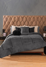 A neatly made bed features the Intima Hogar Cobertor Flannel Extra Suave Estambul in gray, paired with black and gray pillows and a tufted brown headboard. Two nightstands with lamps and framed photos sit against a textured gray wall.