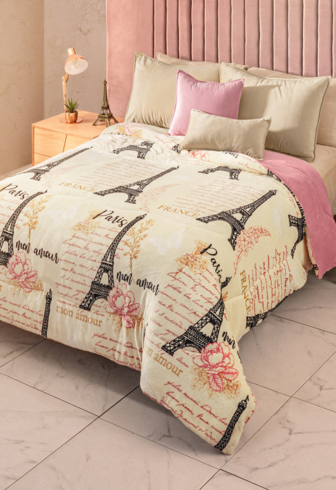 A neatly made bed features the Cobertor Flannel Extra Suave Francia by Intima Hogar, adorned with Eiffel Tower designs, French words, and pink flowers, plus beige and pink pillows, a light pink headboard, and a wooden nightstand with a lamp.