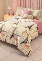 A neatly made bed features the Cobertor Flannel Extra Suave Francia by Intima Hogar, adorned with Eiffel Tower designs, French words, and pink flowers, plus beige and pink pillows, a light pink headboard, and a wooden nightstand with a lamp.