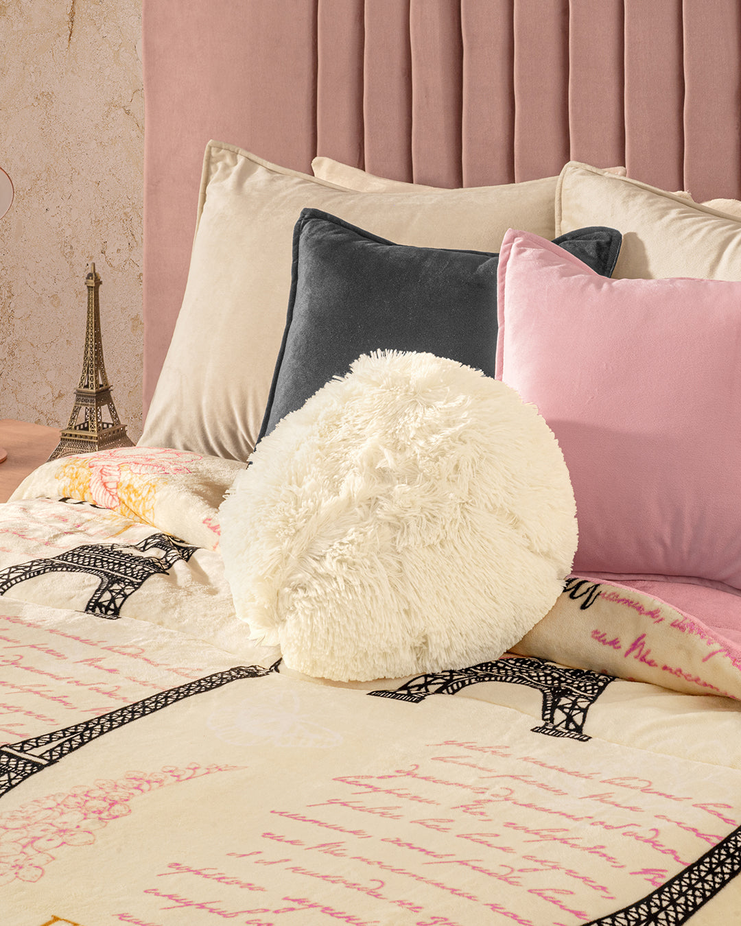 A cozy bed with pink and gray pillows, a round fluffy white pillow, and the Intima Hogar Cobertor Flannel Extra Suave Francia featuring Eiffel Tower designs. A small Eiffel Tower figurine rests on the bedside table by a pink headboard.