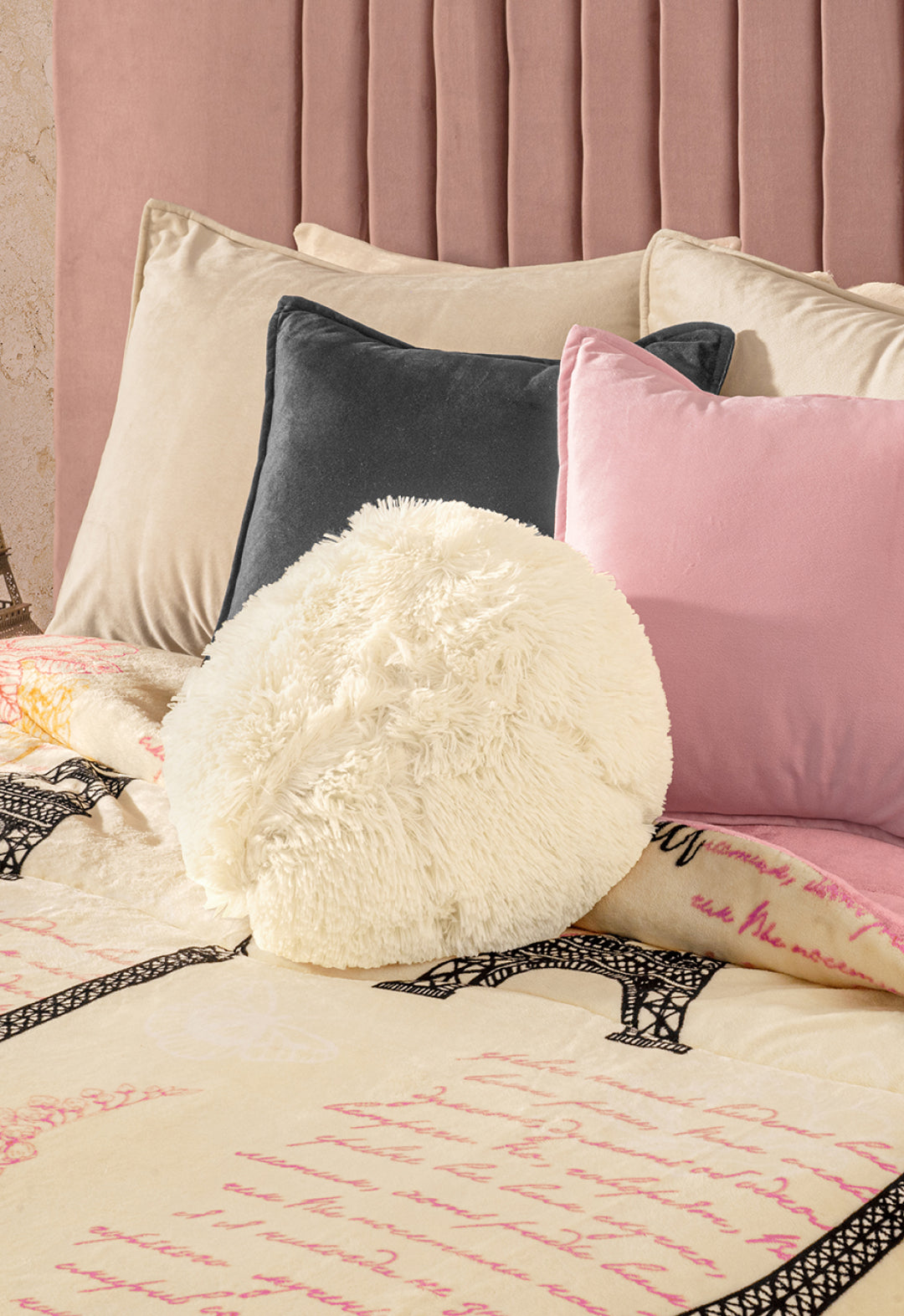 A neatly made bed with beige, black, and pink pillows, a fluffy white round pillow, and the Intima Hogar Cobertor Flannel Extra Suave Francia with Eiffel Tower designs and pink script, set against a padded blush headboard and a romantic touch.