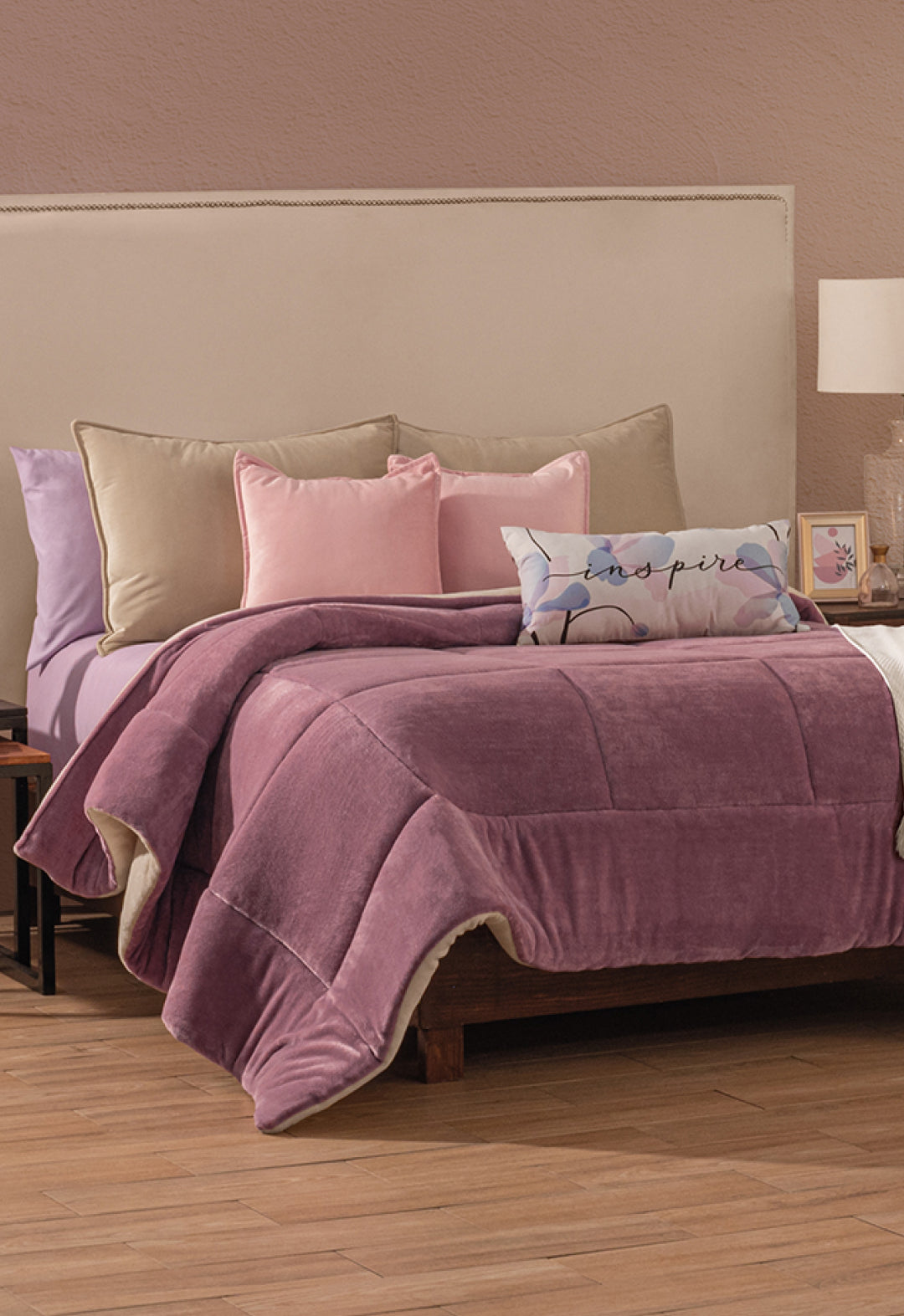 A neatly made bed with the Intima Hogar Cobertor Flannel Extra Suave Italia, beige and pink pillows, and a decorative "inspire" pillow. Wooden floors, neutral walls, and bedside tables create the perfect calidez invierno.