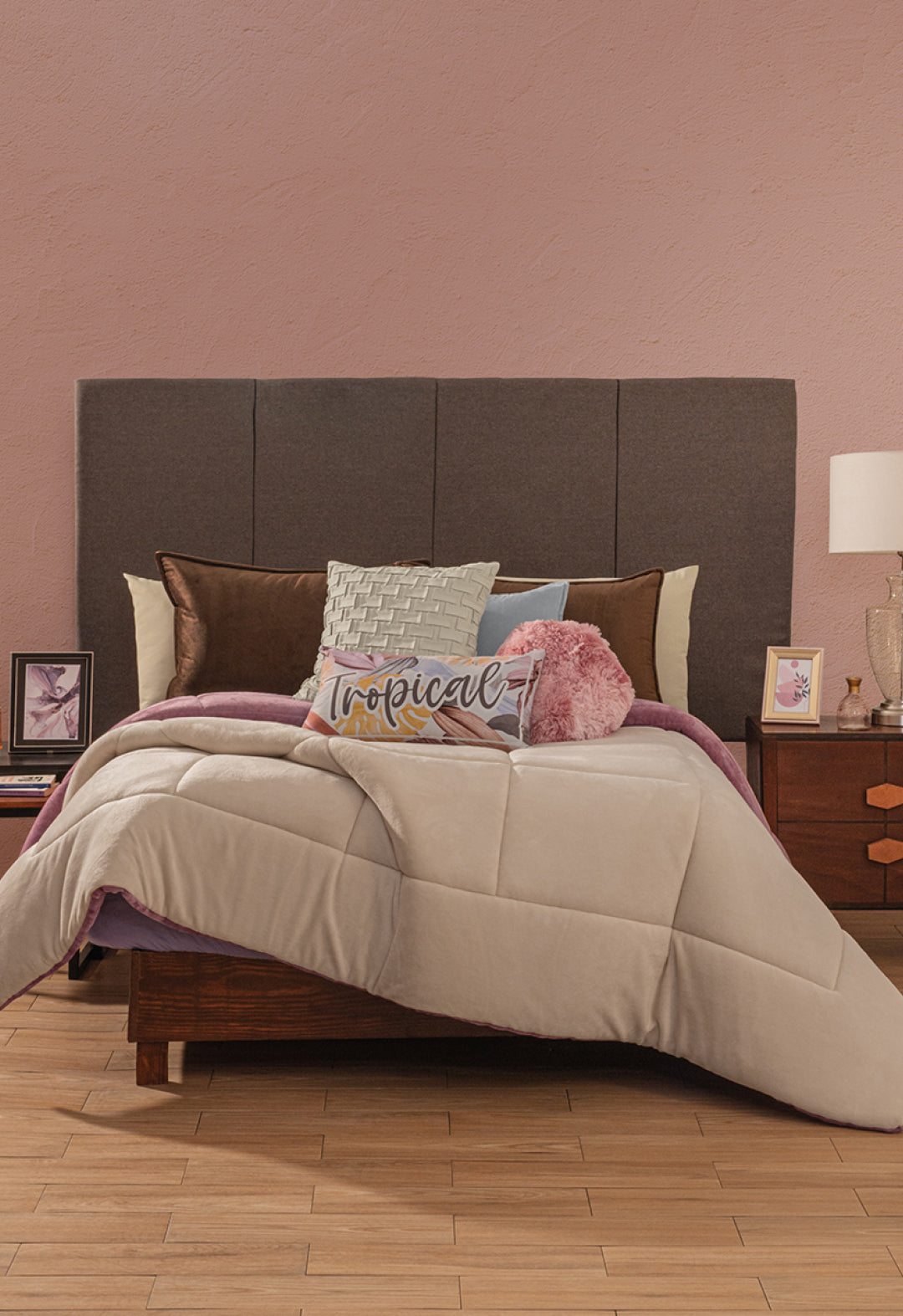 A neatly made bed with pillows, one reading "Tropical," features the Intima Hogar Cobertor Flannel Extra Suave Italia against a pink wall and dark headboard. Two wooden nightstands with lamps, photos, and a vase add cozy modern style.