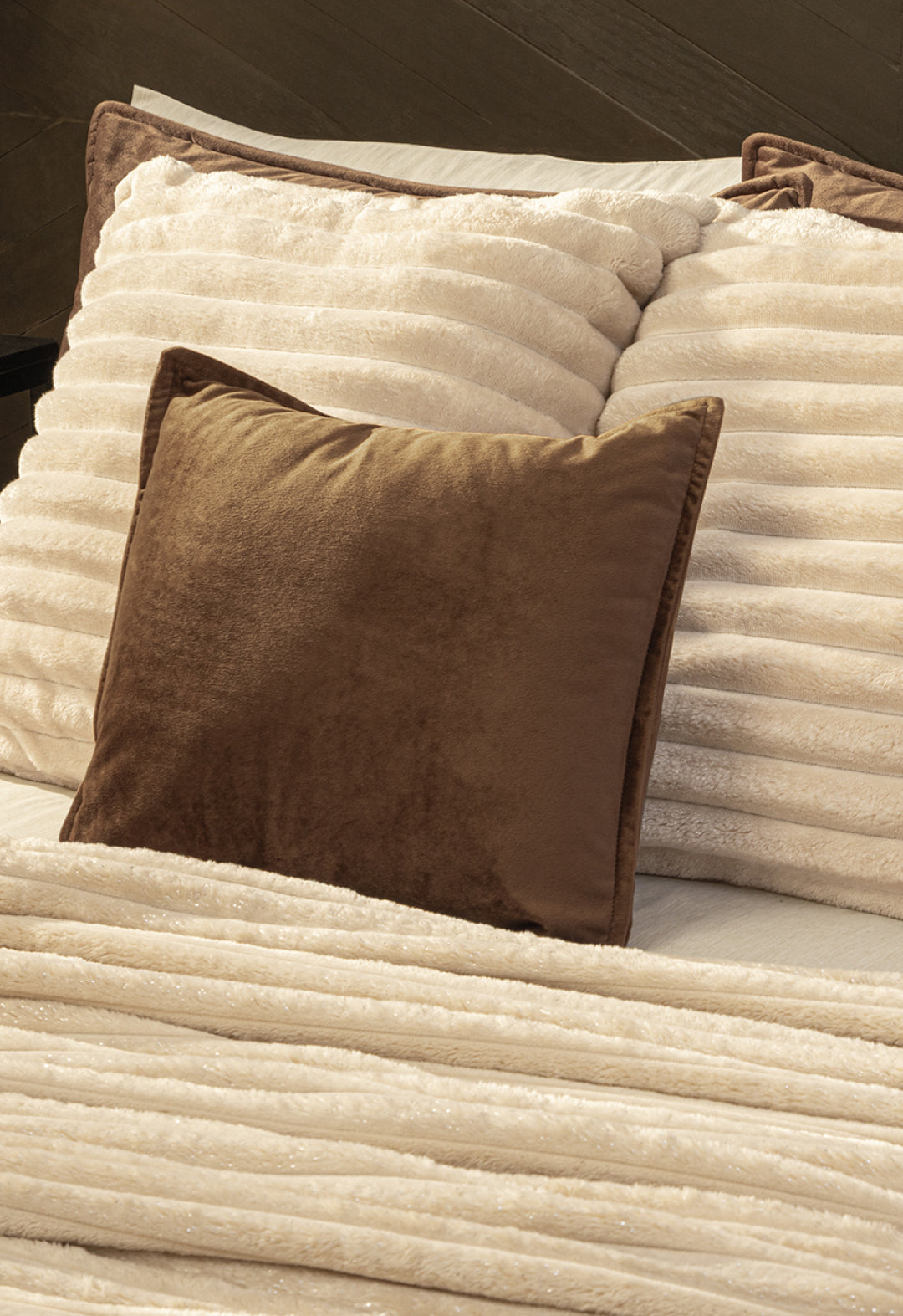 A bed with two cream ribbed pillows and the Cobertor Flannel Ligero Jasper by Intima Hogar in beige, plus a solid brown throw pillow in front for a warm, wash-resistant touch.