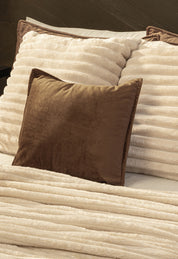 A bed with two cream ribbed pillows and the Cobertor Flannel Ligero Jasper by Intima Hogar in beige, plus a solid brown throw pillow in front for a warm, wash-resistant touch.