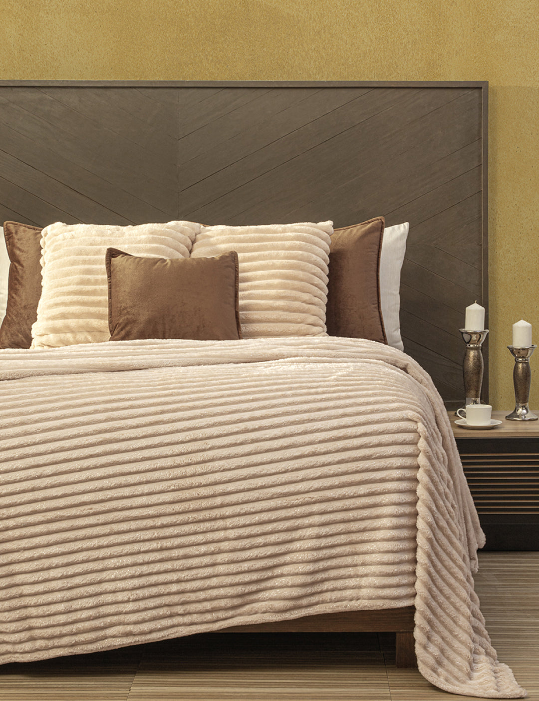 A neatly made bed features the beige Cobertor Flannel Ligero Jasper by Intima Hogar with matching throw pillows and a wooden headboard. Two white candles and cups rest on a dark wood bedside table against a yellow wall.