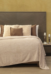 A neatly made bed features the beige Cobertor Flannel Ligero Jasper by Intima Hogar with matching throw pillows and a wooden headboard. Two white candles and cups rest on a dark wood bedside table against a yellow wall.