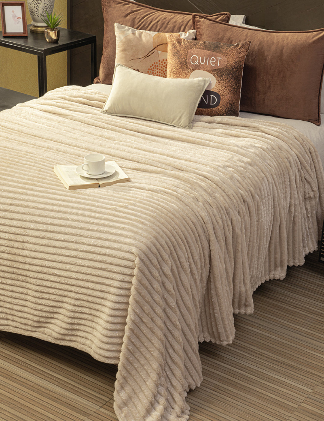 A neatly made bed with the Intima Hogar Cobertor Flannel Ligero Jasper in beige, brown and beige pillows, and a small white tray with a cup and open book. Next to the bed is a side table holding a plant, photo frame, and candle.