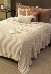A neatly made bed with the Intima Hogar Cobertor Flannel Ligero Jasper in beige, brown and beige pillows, and a small white tray with a cup and open book. Next to the bed is a side table holding a plant, photo frame, and candle.