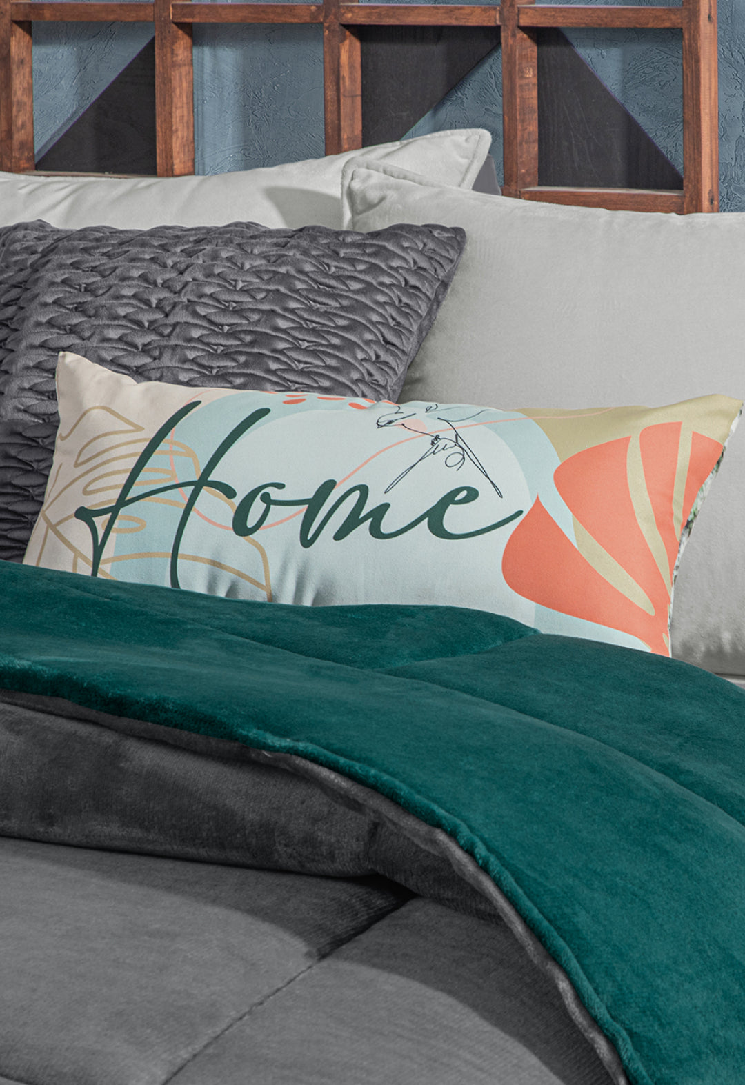 A decorative rectangular pillow with the word Home and abstract patterns rests on a neatly made bed with gray and teal bedding, textured gray pillows, and the Intima Hogar Cobertor Flannel Extra Suave Mediterráneo against a wooden headboard.