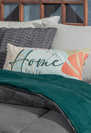 A decorative rectangular pillow with the word Home and abstract patterns rests on a neatly made bed with gray and teal bedding, textured gray pillows, and the Intima Hogar Cobertor Flannel Extra Suave Mediterráneo against a wooden headboard.