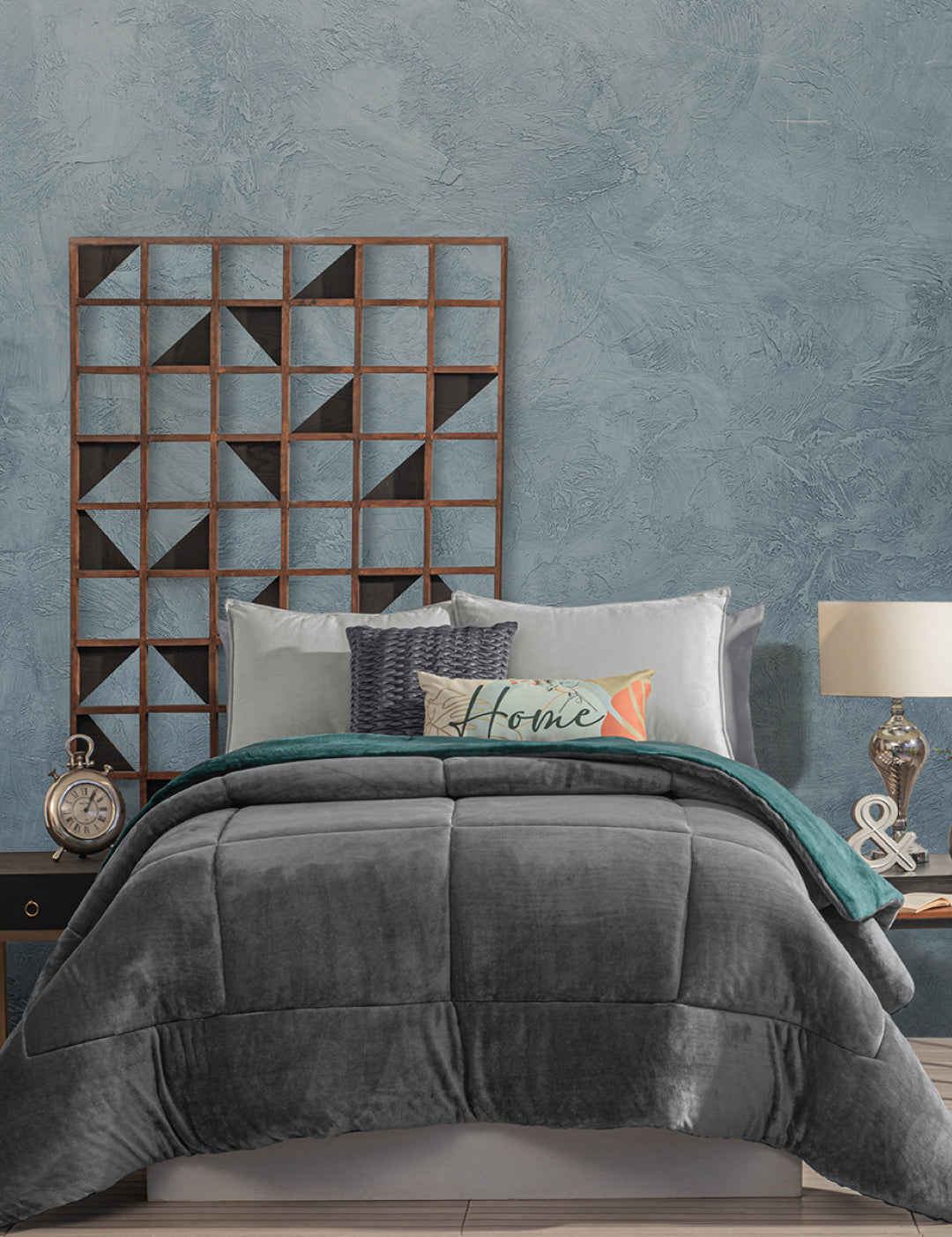A neatly made bed with gray and teal bedding features the Intima Hogar Cobertor Flannel Extra Suave Mediterráneo for a cozy touch, accented by patterned and Home pillows. A wooden geometric wall piece decorates the textured blue wall behind the bed.
