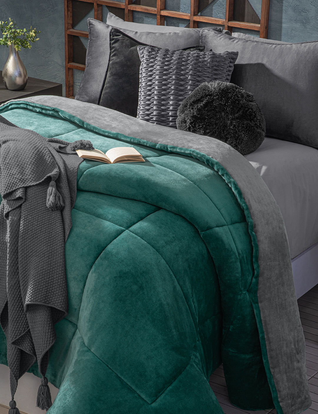 A neatly made bed features the Cobertor Flannel Extra Suave Mediterráneo by Intima Hogar in teal and gray, paired with gray pillows, textured cushions, a tasseled knit throw, and an open book. A flower vase decorates the nearby bedside table.