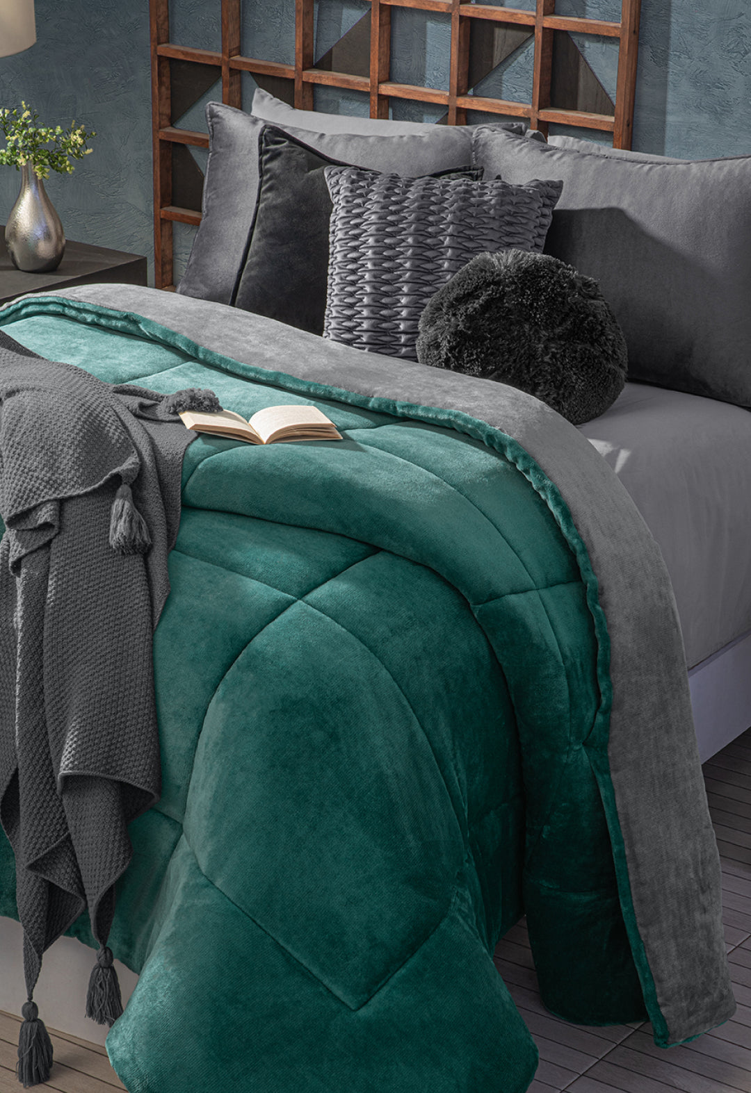 A neatly made bed features the Cobertor Flannel Extra Suave Mediterráneo by Intima Hogar in teal and gray, paired with gray pillows, textured cushions, a tasseled knit throw, and an open book. A flower vase decorates the nearby bedside table.