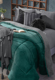 A neatly made bed features the Cobertor Flannel Extra Suave Mediterráneo by Intima Hogar in teal and gray, paired with gray pillows, textured cushions, a tasseled knit throw, and an open book. A flower vase decorates the nearby bedside table.