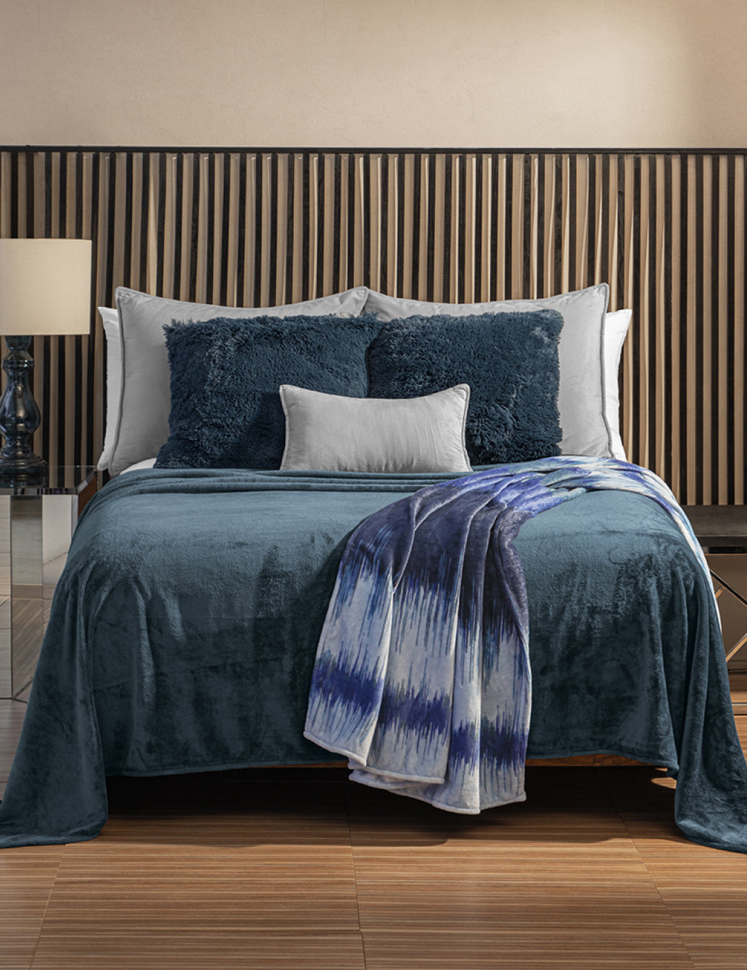 A modern bedroom with a neatly made bed featuring blue and white pillows, an Intima Hogar Cobertor Flannel Ligero Ontario Azul Marino, wooden slat headboard, and a lamp on the nightstand.