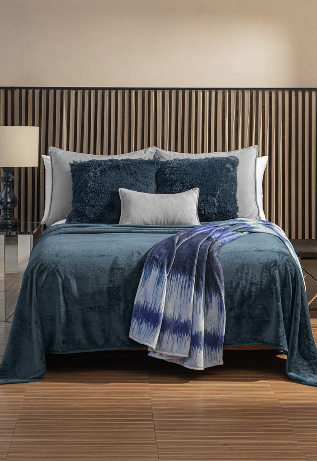 A modern bedroom with a neatly made bed featuring blue and white pillows, an Intima Hogar Cobertor Flannel Ligero Ontario Azul Marino, wooden slat headboard, and a lamp on the nightstand.