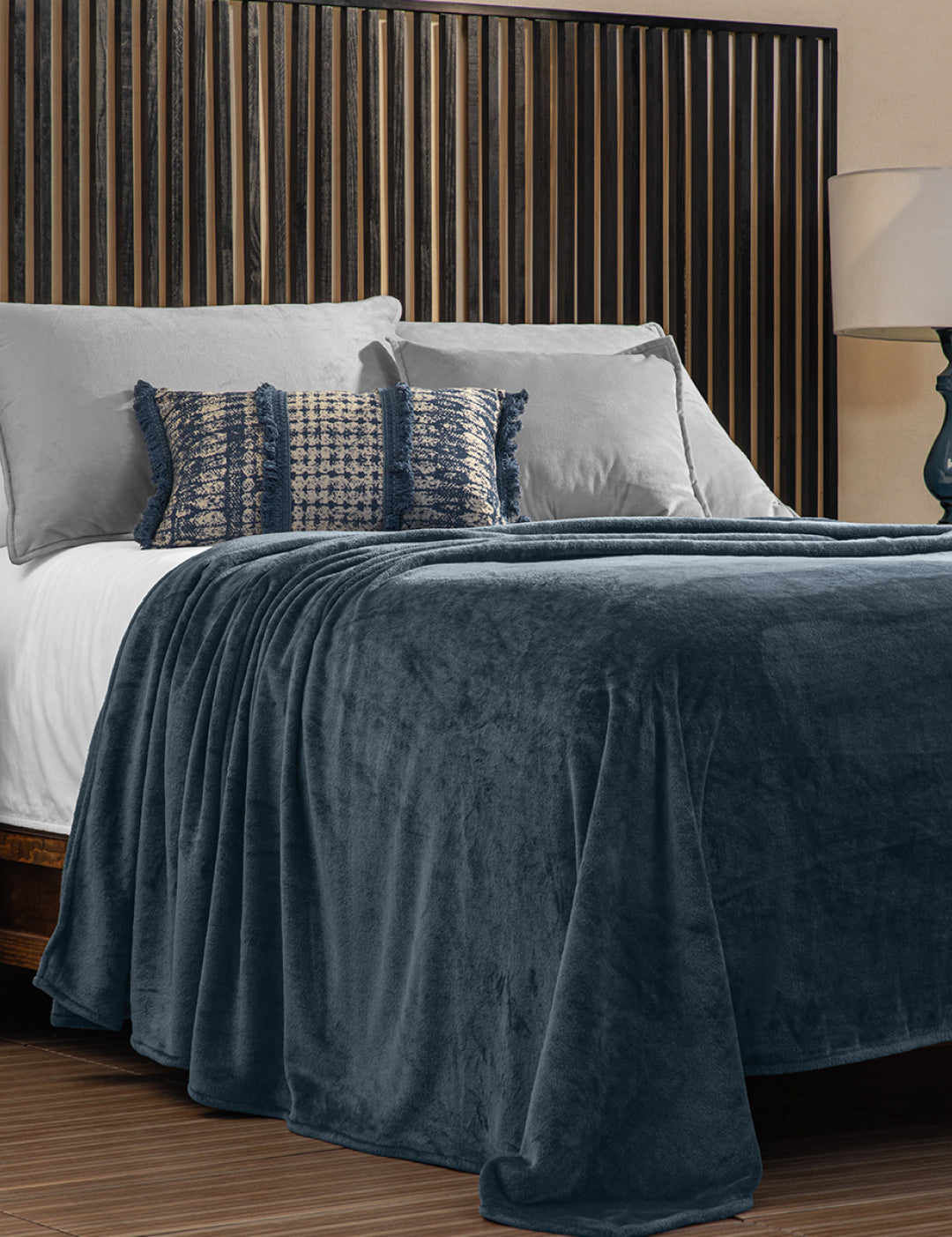 A neatly made bed with gray and textured pillows is covered by the Intima Hogar Cobertor Flannel Ligero Ontario Azul Marino, set before a vertical wooden slat headboard. A lamp sits on the bedside table.
