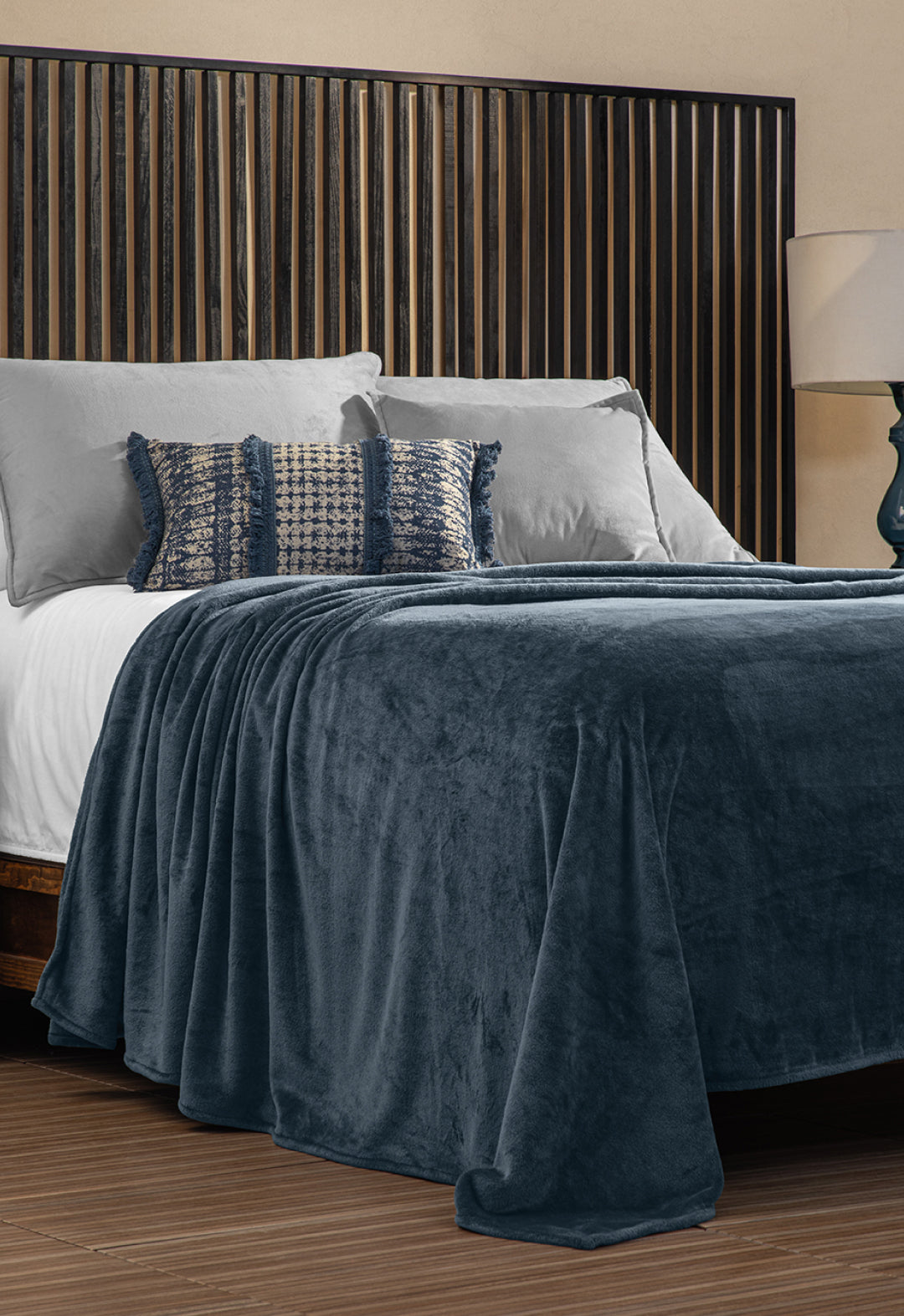 A neatly made bed with gray and textured pillows is covered by the Intima Hogar Cobertor Flannel Ligero Ontario Azul Marino, set before a vertical wooden slat headboard. A lamp sits on the bedside table.