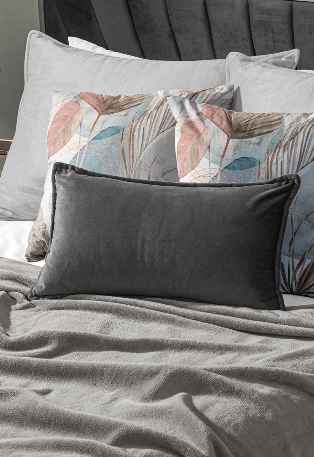 A neatly made bed with a dark gray upholstered headboard, two light gray pillows, a pink and blue leaf-patterned pillow, a dark gray velvet pillow, and the Intima Hogar Cobertor Flannel Ligero Ontario Gris.