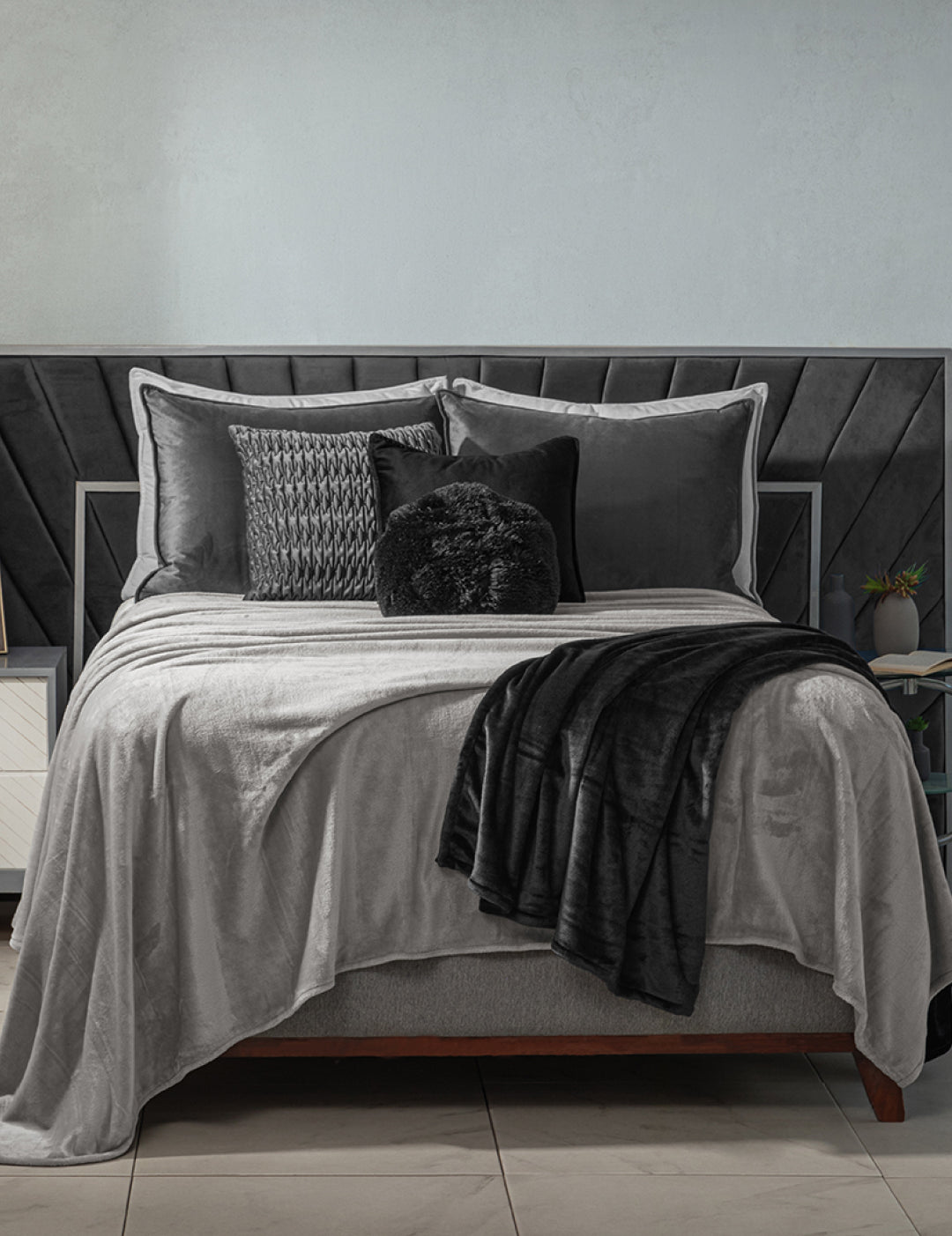A neatly made bed features the Intima Hogar Cobertor Flannel Ligero Ontario Gris with gray and black bedding, decorative pillows, and a black textured headboard. A white nightstand and a plant are beside the bed.