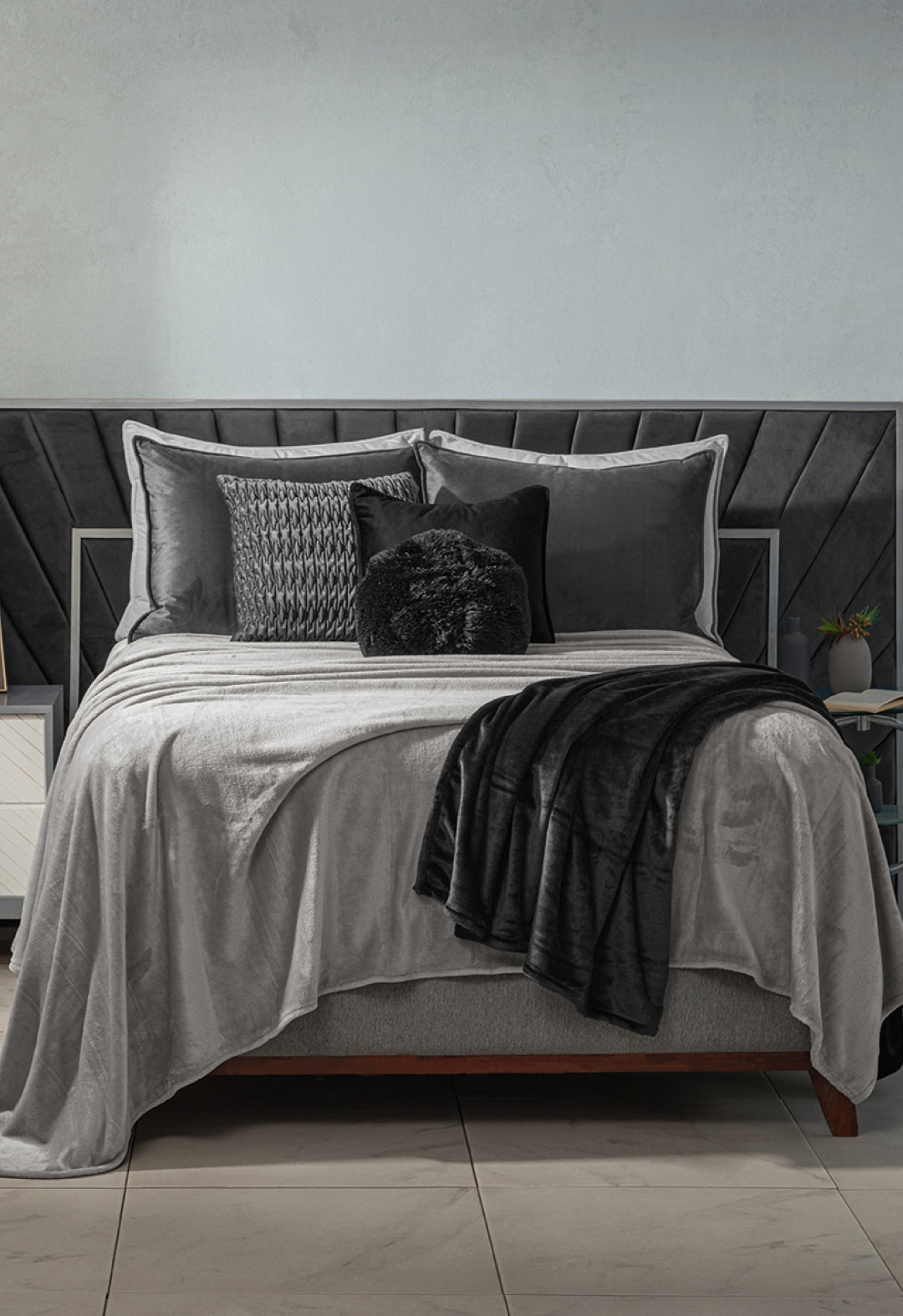 A neatly made bed features the Intima Hogar Cobertor Flannel Ligero Ontario Gris with gray and black bedding, decorative pillows, and a black textured headboard. A white nightstand and a plant are beside the bed.