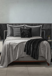 A neatly made bed features the Intima Hogar Cobertor Flannel Ligero Ontario Gris with gray and black bedding, decorative pillows, and a black textured headboard. A white nightstand and a plant are beside the bed.