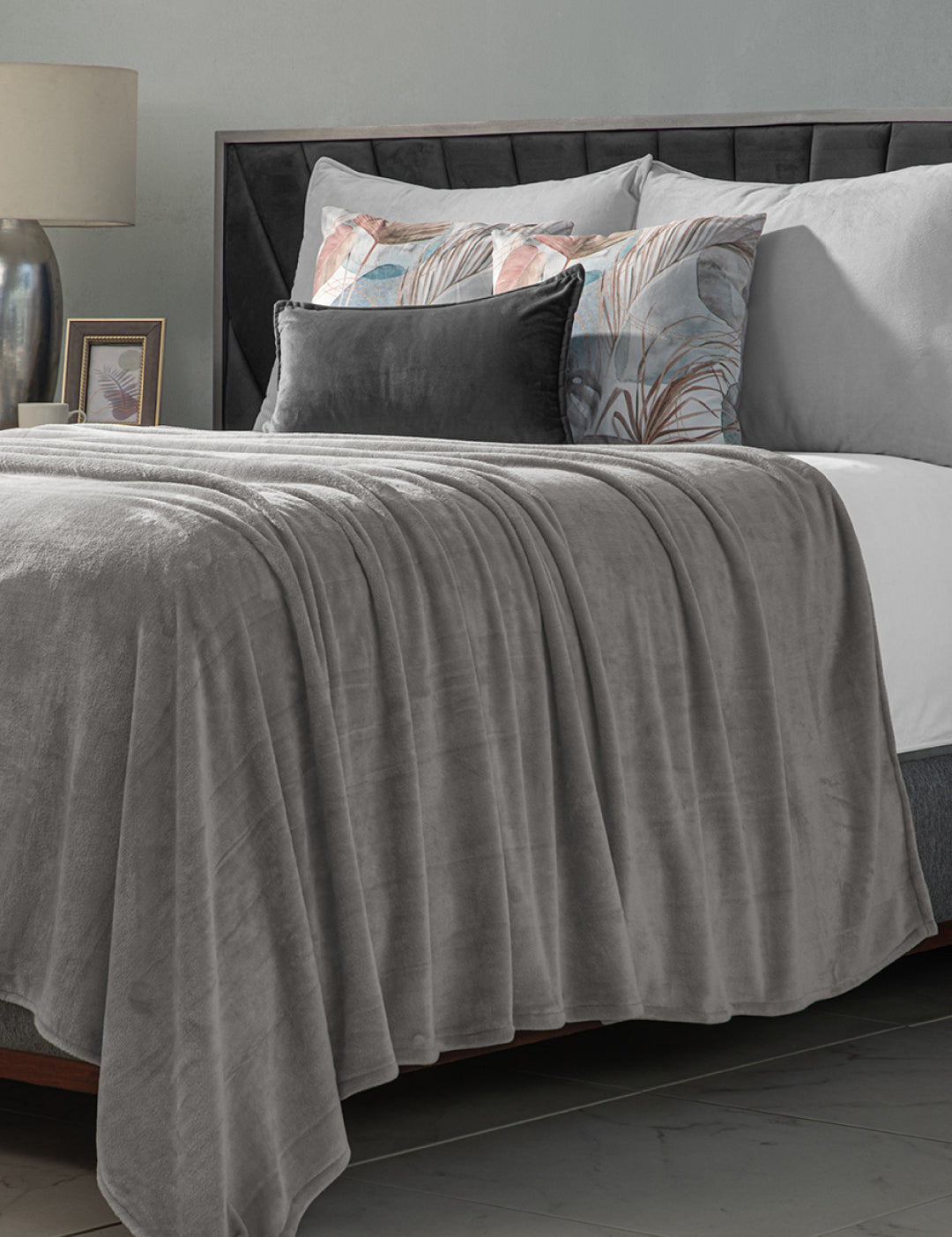 A neatly made bed with the Cobertor Flannel Ligero Ontario Gris by Intima Hogar, gray and pastel decorative pillows, a bedside table with a lamp and photo frame, set against a gray wall in the background.