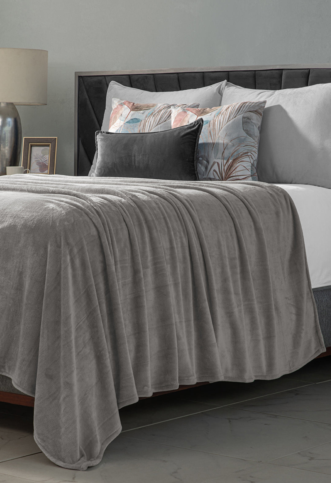 A neatly made bed with the Cobertor Flannel Ligero Ontario Gris by Intima Hogar, gray and pastel decorative pillows, a bedside table with a lamp and photo frame, set against a gray wall in the background.