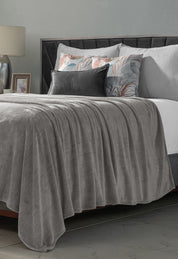A neatly made bed with the Cobertor Flannel Ligero Ontario Gris by Intima Hogar, gray and pastel decorative pillows, a bedside table with a lamp and photo frame, set against a gray wall in the background.