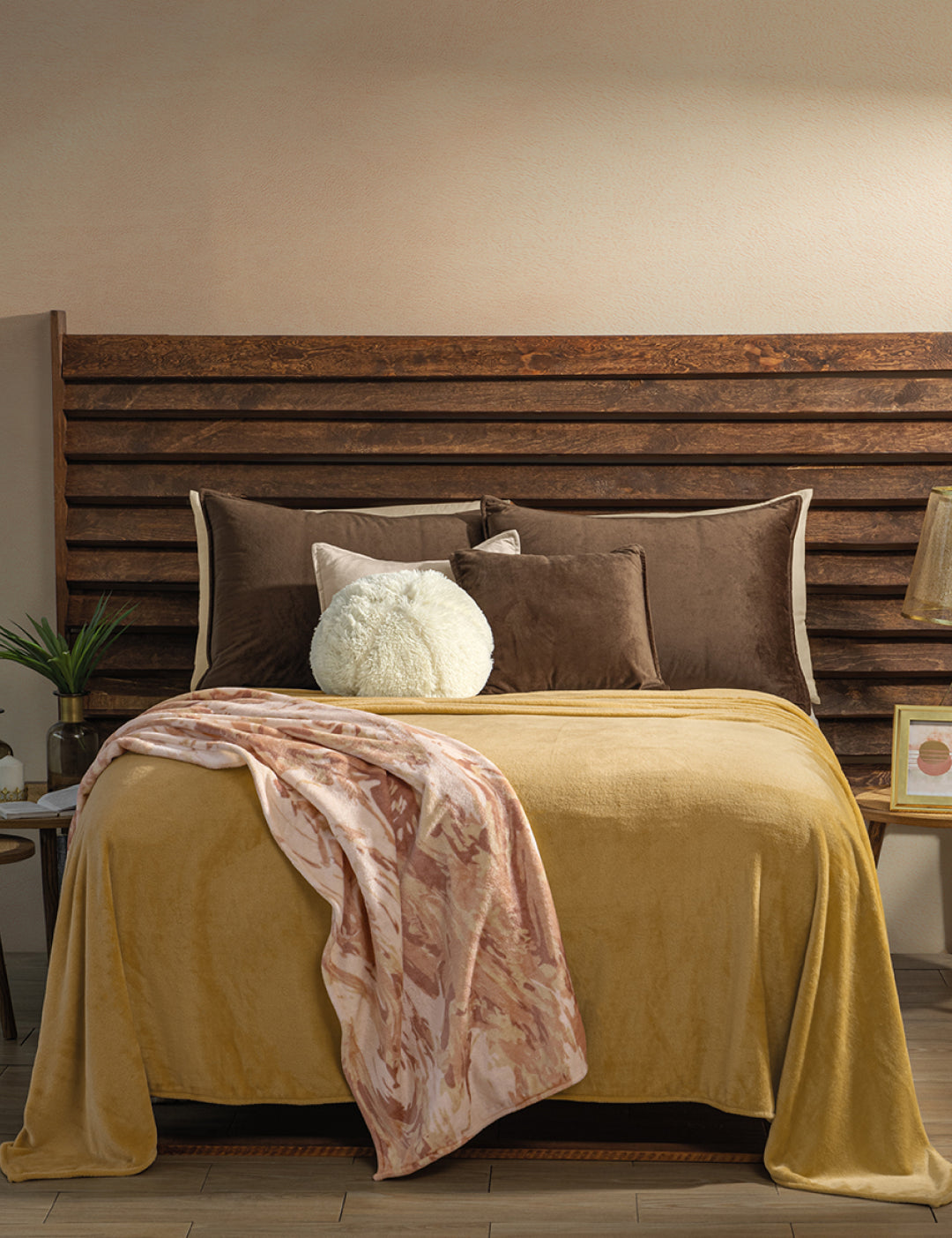 A neatly made bed with brown and mustard yellow bedding, accented by brown, white, and taupe pillows. The Cobertor Flannel Ligero Ontario Oro by Intima Hogar is draped over the foot, with nightstands and decor on each side.