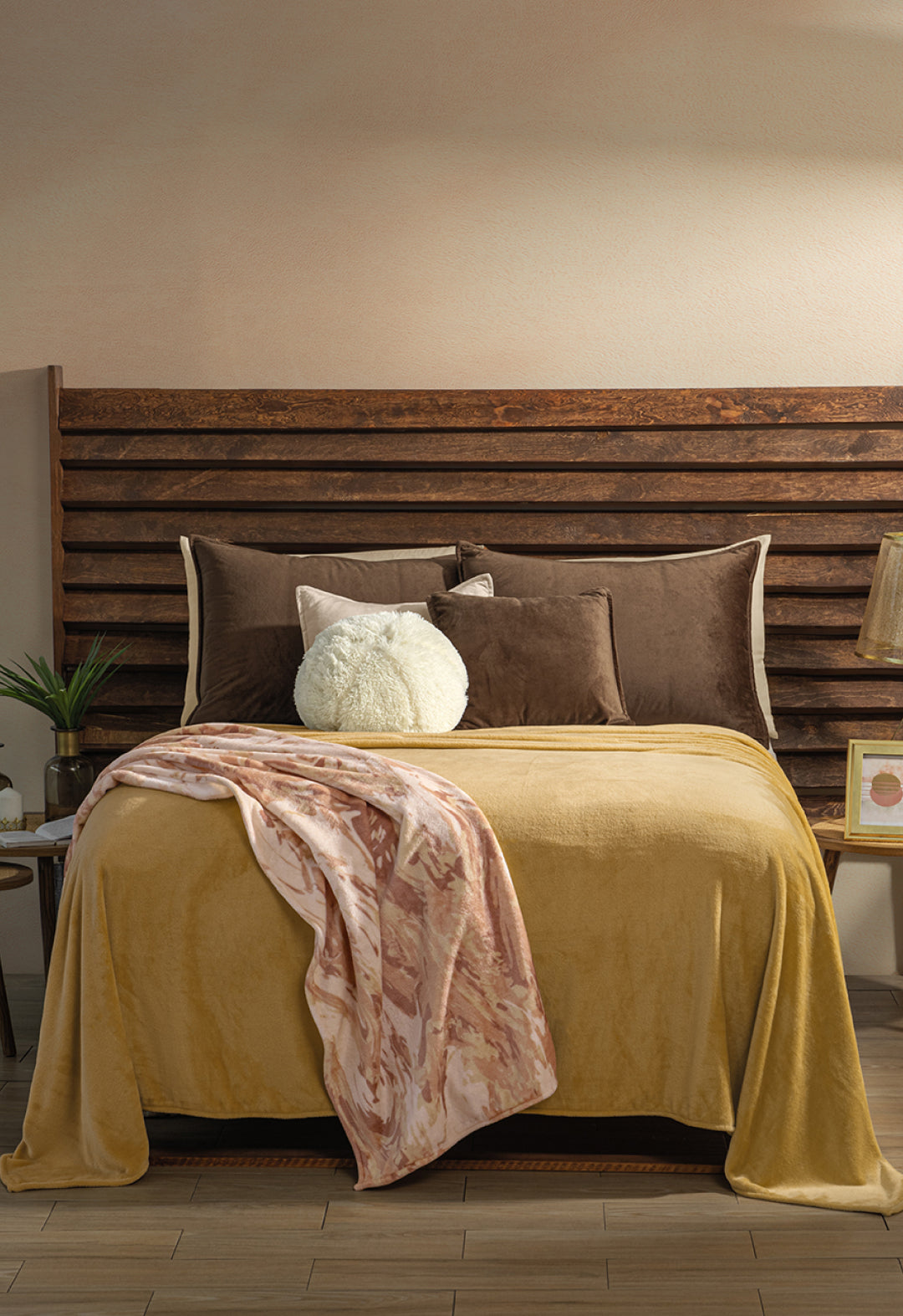A neatly made bed with brown and mustard yellow bedding, accented by brown, white, and taupe pillows. The Cobertor Flannel Ligero Ontario Oro by Intima Hogar is draped over the foot, with nightstands and decor on each side.