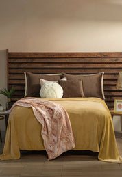 A neatly made bed with brown and mustard yellow bedding, accented by brown, white, and taupe pillows. The Cobertor Flannel Ligero Ontario Oro by Intima Hogar is draped over the foot, with nightstands and decor on each side.