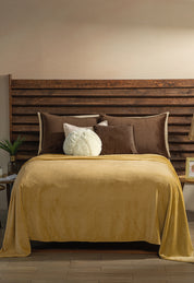 A neatly made bed with a wooden slatted headboard features the Cobertor Flannel Ligero Ontario Oro by Intima Hogar, brown and beige pillows, a round white pillow, and bedside tables with small plants and picture frames against a light wall.