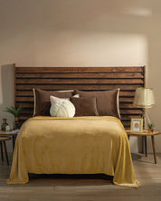 A cozy habitación with a wooden slatted headboard, a cama covered in a yellow blanket and Intima Hogar’s Funda de Almohada Ensueño Café pillows, round side tables with plants, a lamp, framed photo, and a small candle on the wooden floor.