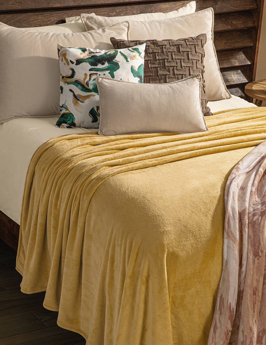 A neatly made bed with an Intima Hogar Cobertor Flannel Ligero Ontario Oro in soft yellow, beige sheets, and four decorative pillows: green and gold patterned, brown woven, solid beige, and a rectangular light pillow.