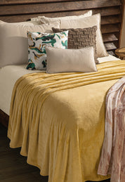 A neatly made bed with an Intima Hogar Cobertor Flannel Ligero Ontario Oro in soft yellow, beige sheets, and four decorative pillows: green and gold patterned, brown woven, solid beige, and a rectangular light pillow.