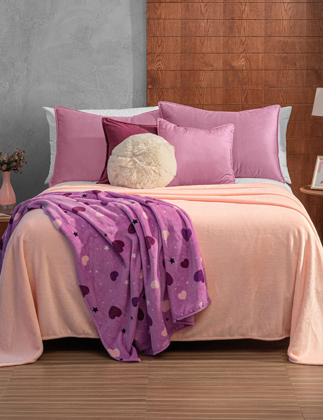 A neatly made bed with pink and purple pillows, a fluffy round cushion, and a peach blanket is topped with the Intima Hogar Cobertor Flannel Ligero Ontario Rosa. The room has a wood-paneled wall and a small bedside table with a vase.