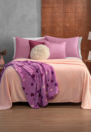 A neatly made bed with pink and purple pillows, a fluffy round cushion, and a peach blanket is topped with the Intima Hogar Cobertor Flannel Ligero Ontario Rosa. The room has a wood-paneled wall and a small bedside table with a vase.