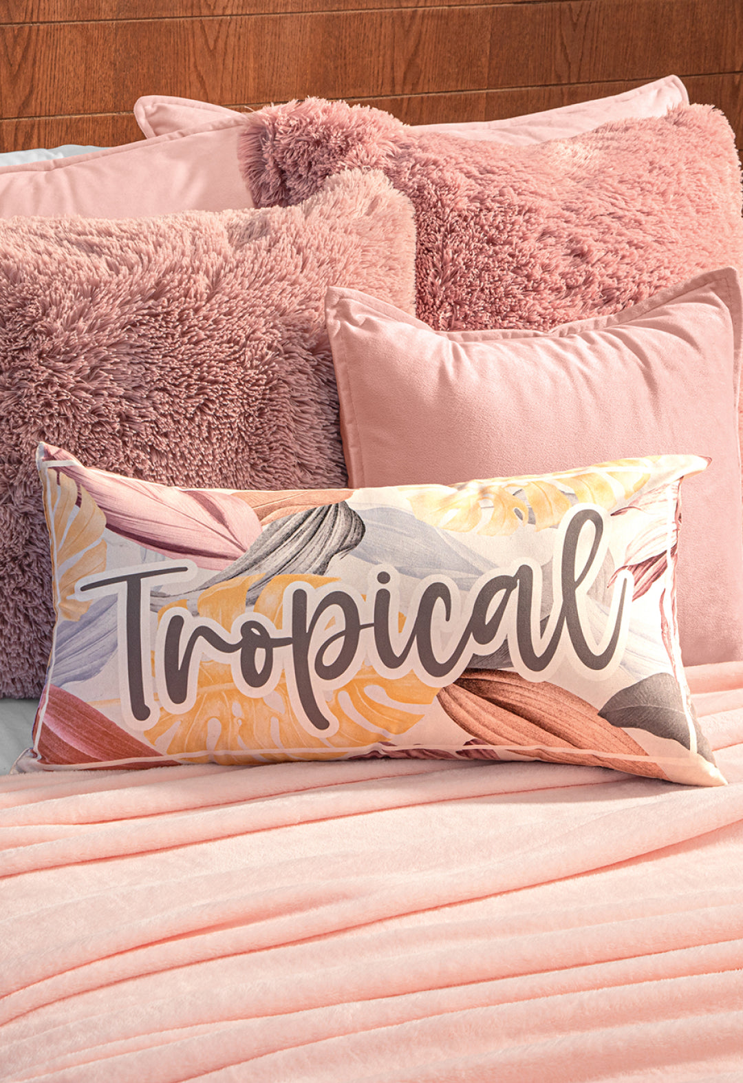A bed features the Intima Hogar Cobertor Flannel Ligero Ontario Rosa, paired with soft, textured pink pillows and a rectangular accent pillow decorated with pastel leaves and "Tropical" boldly scripted.