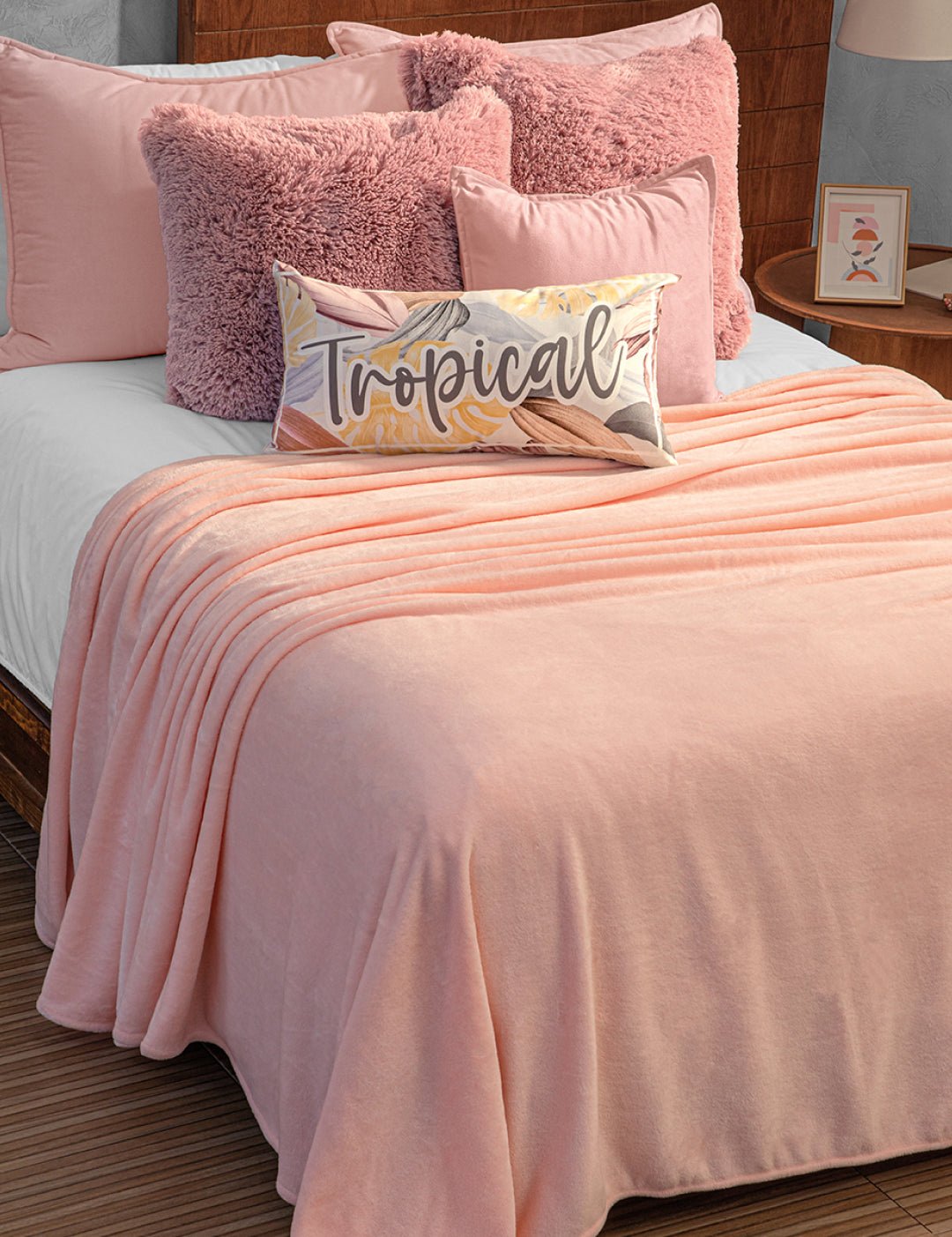 A neatly made bed with the Intima Hogar Cobertor Flannel Ligero Ontario Rosa, pink pillows, fluffy cushions, and a "Tropical" script pillow. A wooden headboard and bedside lamp complete the perfect comfort night.
