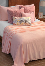 A neatly made bed with the Intima Hogar Cobertor Flannel Ligero Ontario Rosa, pink pillows, fluffy cushions, and a "Tropical" script pillow. A wooden headboard and bedside lamp complete the perfect comfort night.