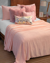 A neatly made bed features Cojines de Lujo Liverpool by Intima Hogar, including a pink blanket, pink pillows, a decorative Tropical pillow, and a wooden headboard next to a lamp and small nightstand.